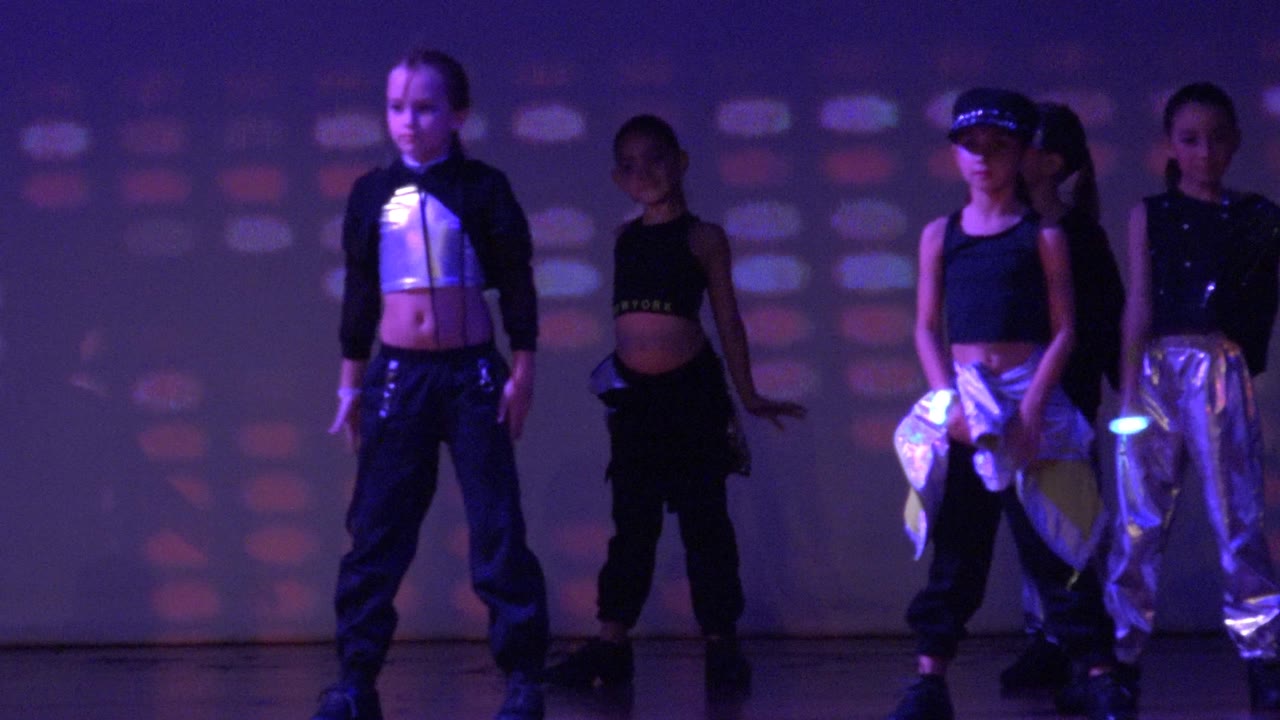 Dancers stand in a staggered formation with glowing wristbands and colored wall projections.