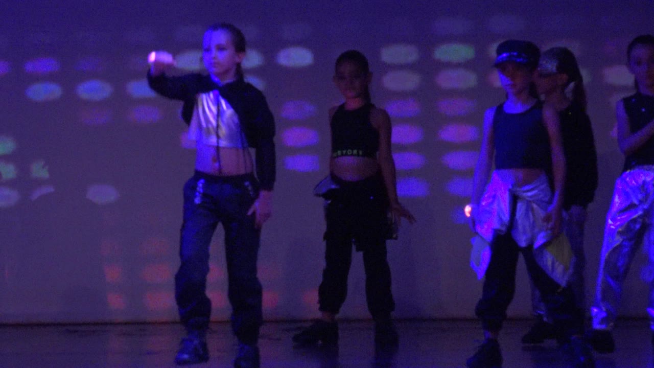 Teen dancers in San Jose Del Cabo BCS Mexico shift through choreography, with a front dancer in a silver top and others partly silhouetted by blue and purple stage light.