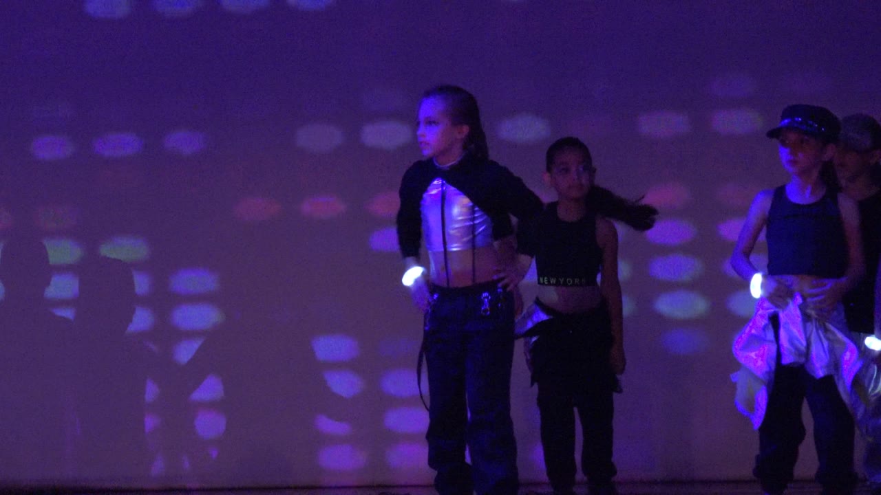 Children in dark dance outfits stand on a stage in San Jose del Cabo, Mexico, with purple light patterns on the rear wall and glowing wrist accessories visible.