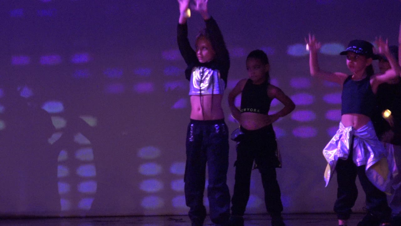 Children dancing with arms raised against a purple lit stage backdrop.