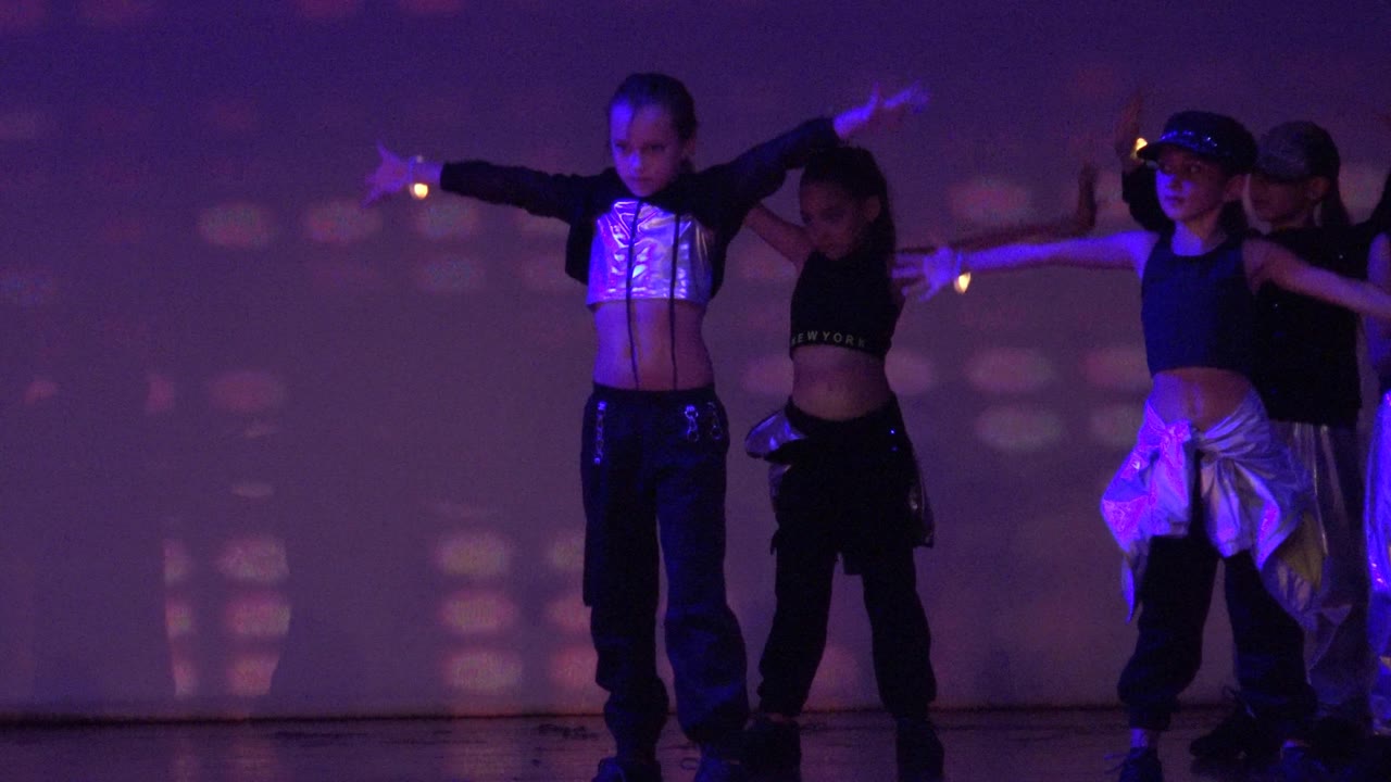 Children in dark outfits hold wide arm positions during a stage dance performance in San Jose del Cabo, Mexico, with colored light patterns behind them.