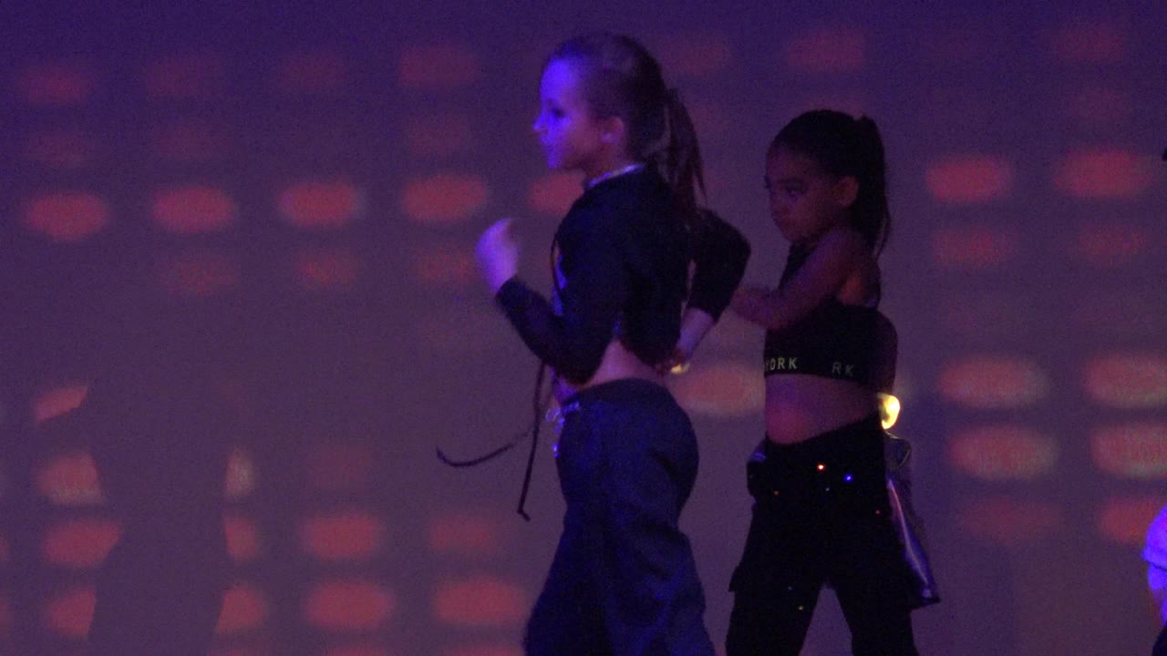 Two girls dance in San Jose Del Cabo BCS Mexico, with one wearing a dark cap as purple, green, and red projected shapes fill the background.