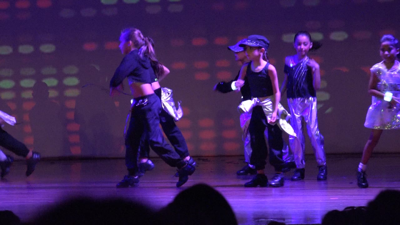 Children perform on a purple-lit stage in San Jose del Cabo, Mexico, with one dancer crossing left and others spaced across the background.