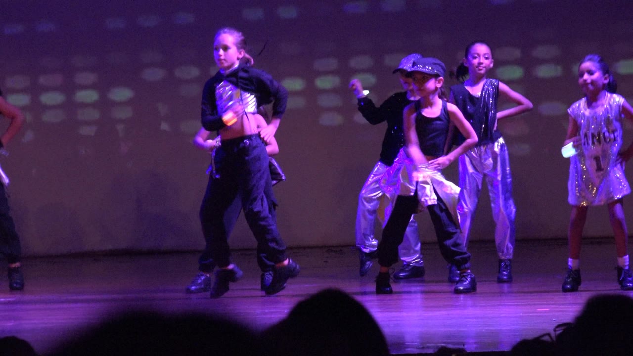 Young dancers in shiny black and silver outfits perform on a purple-lit stage in San Jose Del Cabo BCS Mexico, with audience silhouettes along the bottom edge.