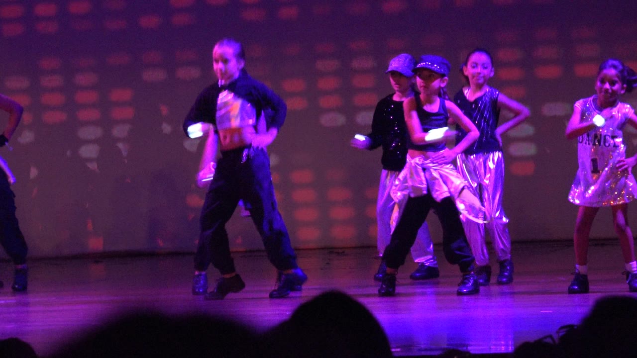 Youth dance group shifts positions under colored stage lights.