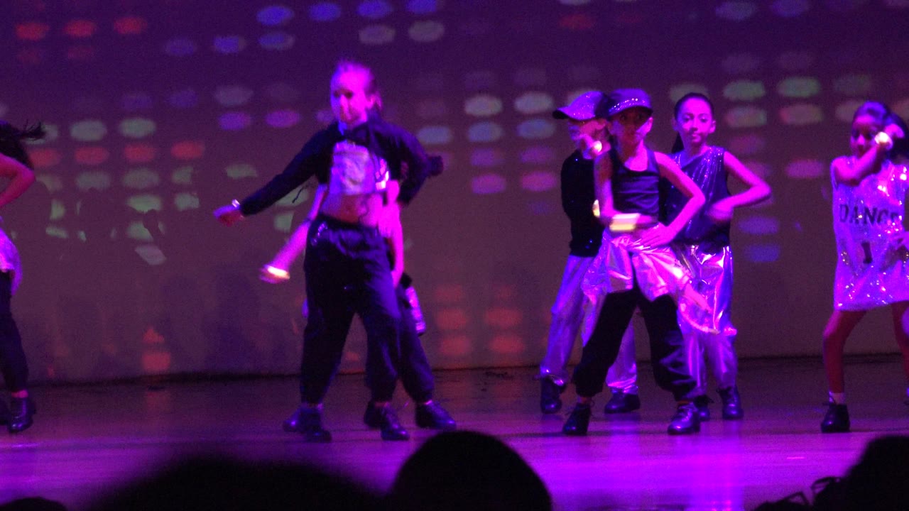 Several children dance in formation on the San Jose Del Cabo BCS Mexico stage, including performers in black, silver, and gold outfits beneath magenta-purple lighting.