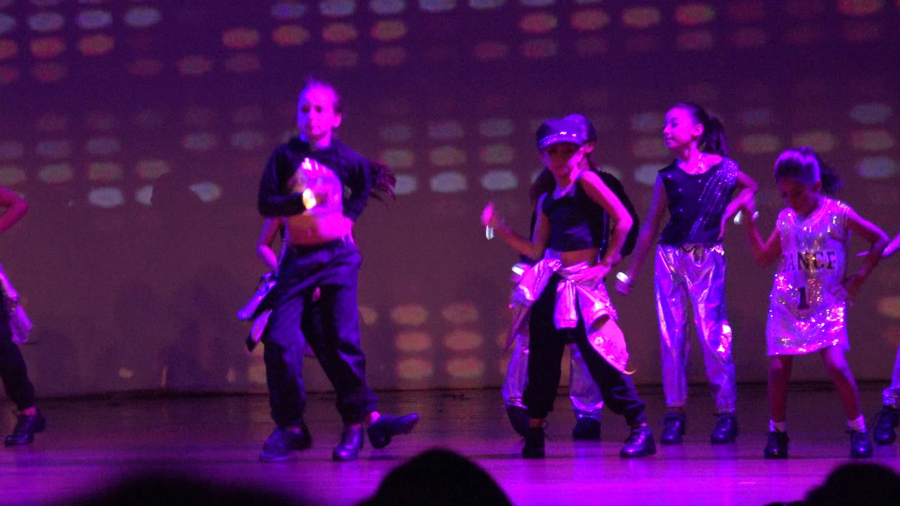 Children in shiny and black dancewear perform in a coordinated line on a San Jose Del Cabo BCS Mexico stage, with purple light and multicolored dots behind them.