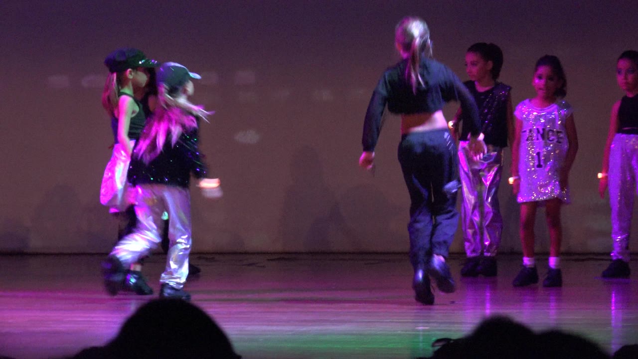 Several children stand and move on the right side of the San Jose del Cabo, Mexico stage, with pink and green light reflecting on shiny costumes.