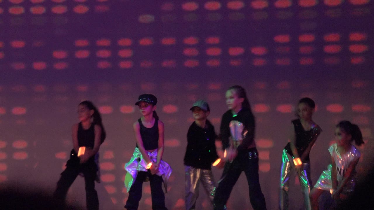 Children in shiny outfits and dark clothing dance with glowing props.
