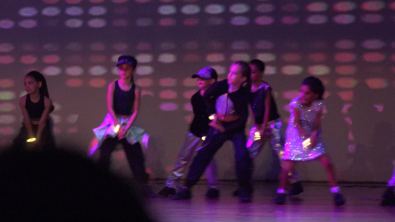 A line of child performers in San Jose del Cabo, Mexico stands across the stage with glow sticks or small light props and a grid-like colored projection behind them.