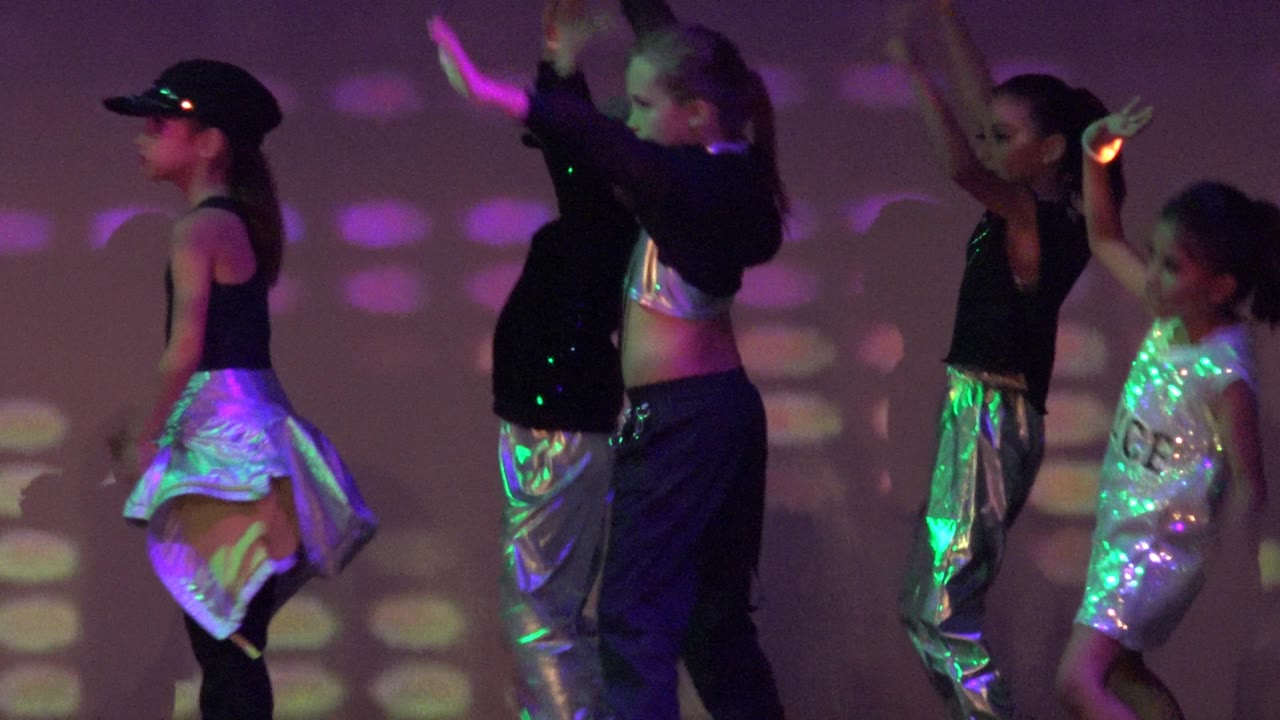 Children dance with raised arms and glow sticks under colored stage lights.