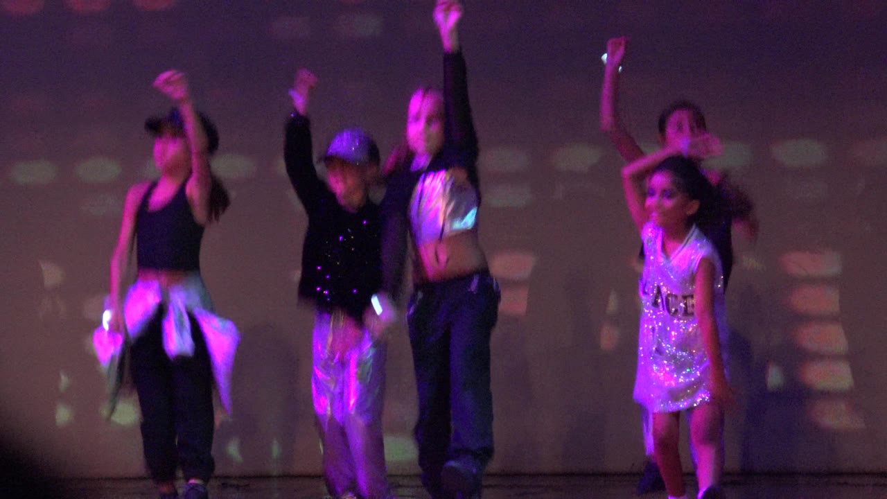 Several children dance in staggered positions on a stage in San Jose Del Cabo BCS Mexico, with glitter costumes catching green and magenta light.