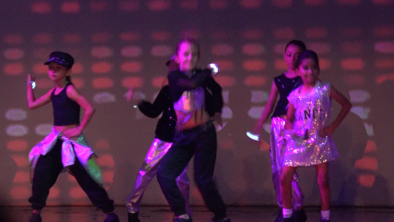 Children perform a synchronized dance on a stage in San Jose Del Cabo, Mexico, with dark outfits, shiny silver costume pieces, and purple-red lighting on the backdrop.