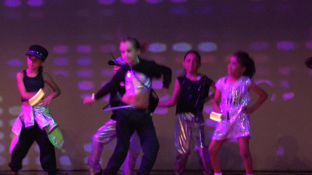 Children perform on a stage in San Jose del Cabo BCS Mexico, wearing shiny dance outfits and holding glowing accessories under purple and pink patterned lighting.