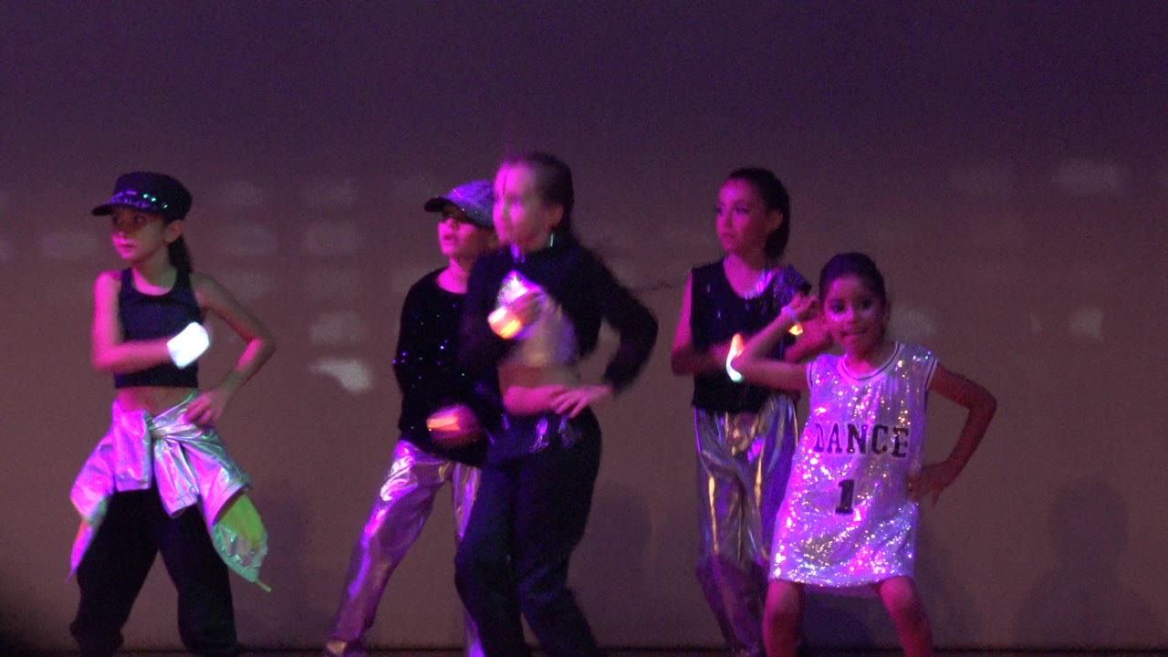 Several children move through the routine on a stage in San Jose del Cabo BCS Mexico, with glowing hand accessories and pink-purple light patterns behind them.