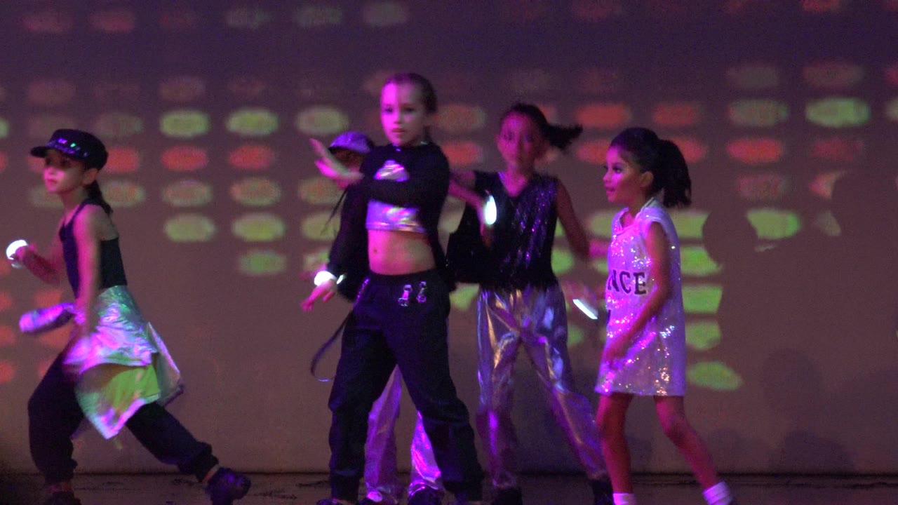 Children perform on a stage in San Jose del Cabo, Mexico, with glow sticks, shiny costumes, and purple-green projected light behind them at the start of the clip.