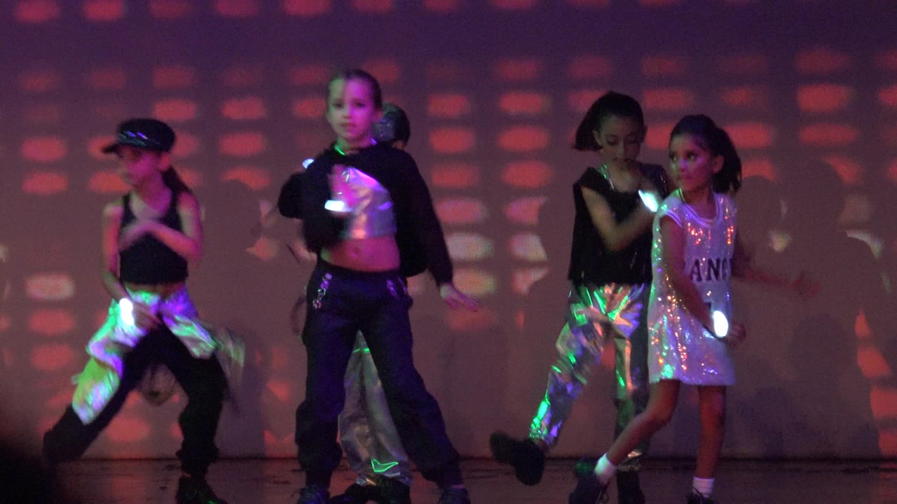 Children move across the stage with glow sticks under multicolored lights.