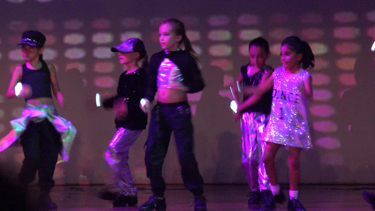 Young dancers gather near center stage in San Jose del Cabo, Mexico, with reflective silver clothing and a white sequined dress catching purple and green light.