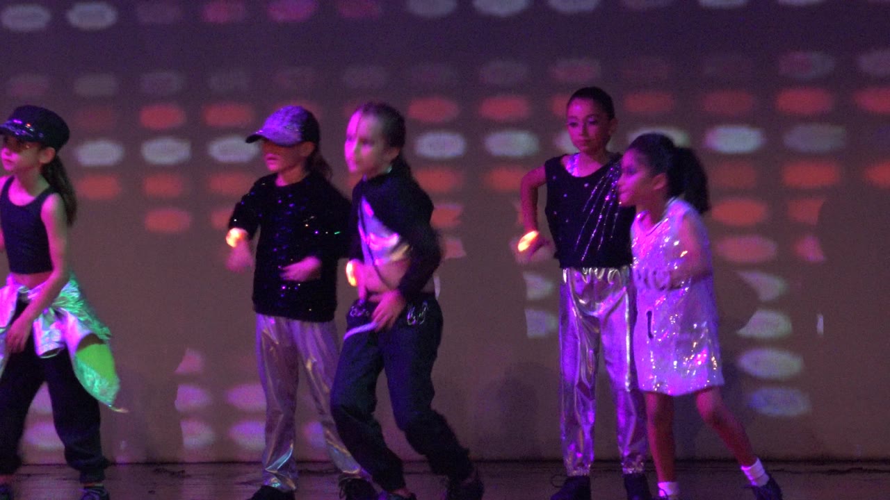 Children perform a dance routine on a stage in San Jose Del Cabo BCS Mexico, with shiny black and silver outfits and purple-red light patterns behind them.