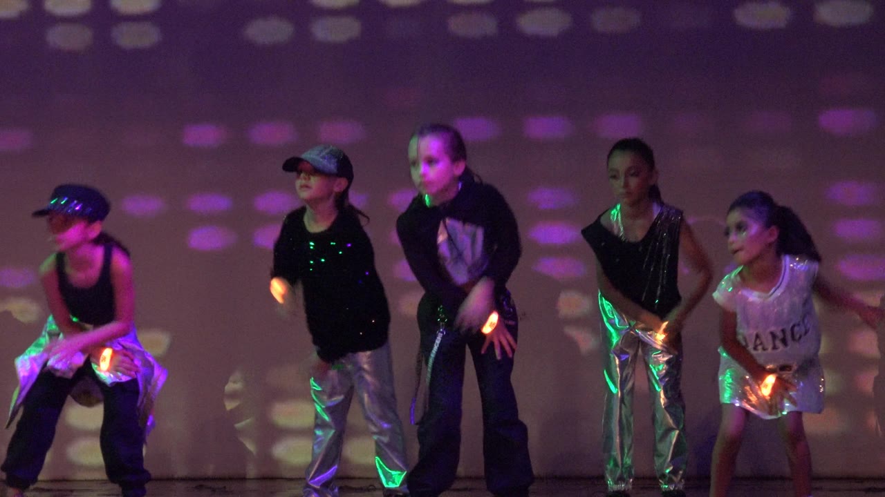 Children hold dance positions on stage with green and purple light reflections.