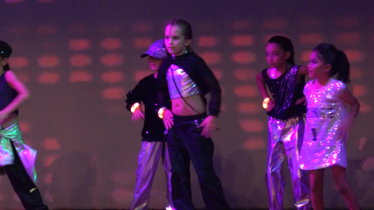 The dance group shifts into a lower formation on stage in San Jose Del Cabo BCS Mexico, with several children bending forward and colored light patterns behind them.