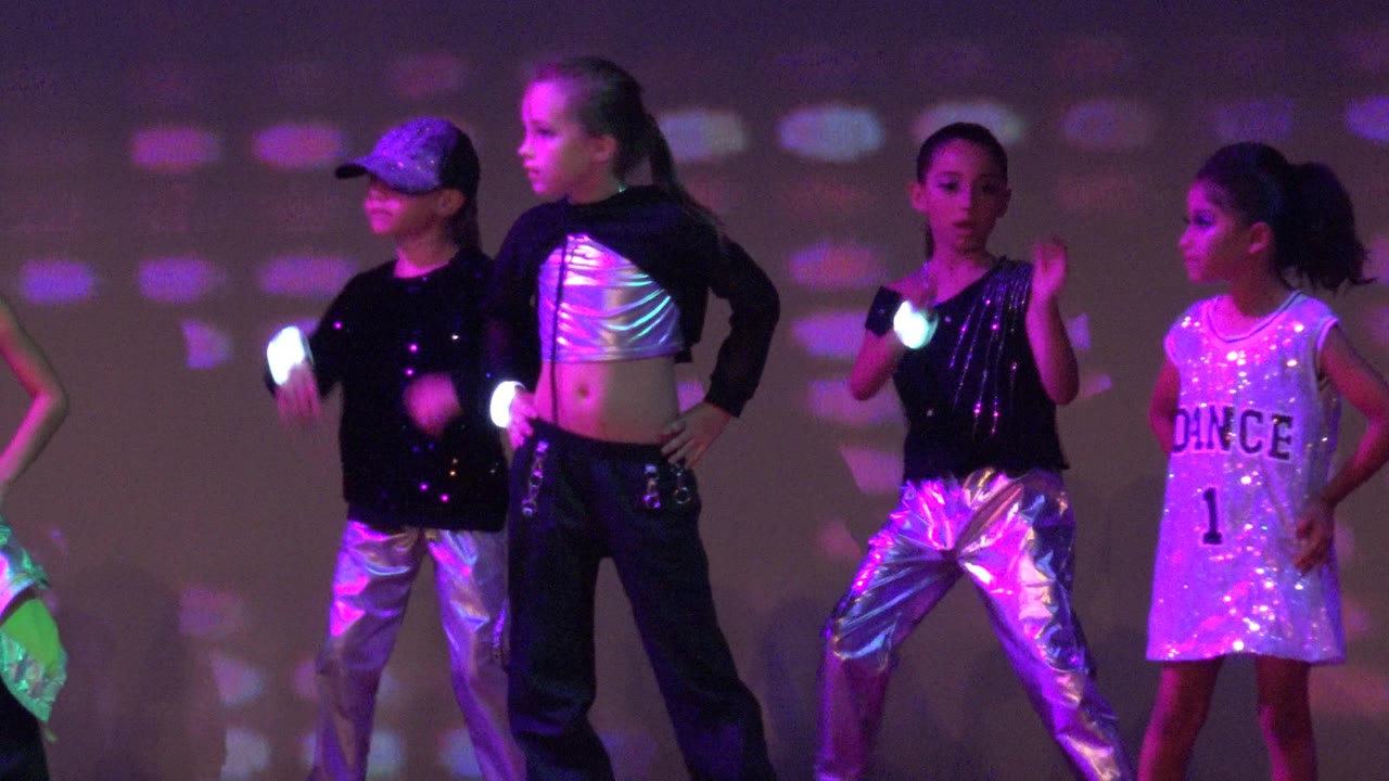 Children in black, silver, and white dance outfits stand across a stage in San Jose del Cabo BCS Mexico as purple and green light patterns cover the backdrop.