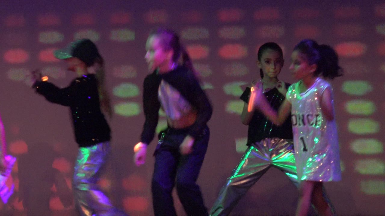 Children dancing on stage in coordinated black, silver, and white outfits.