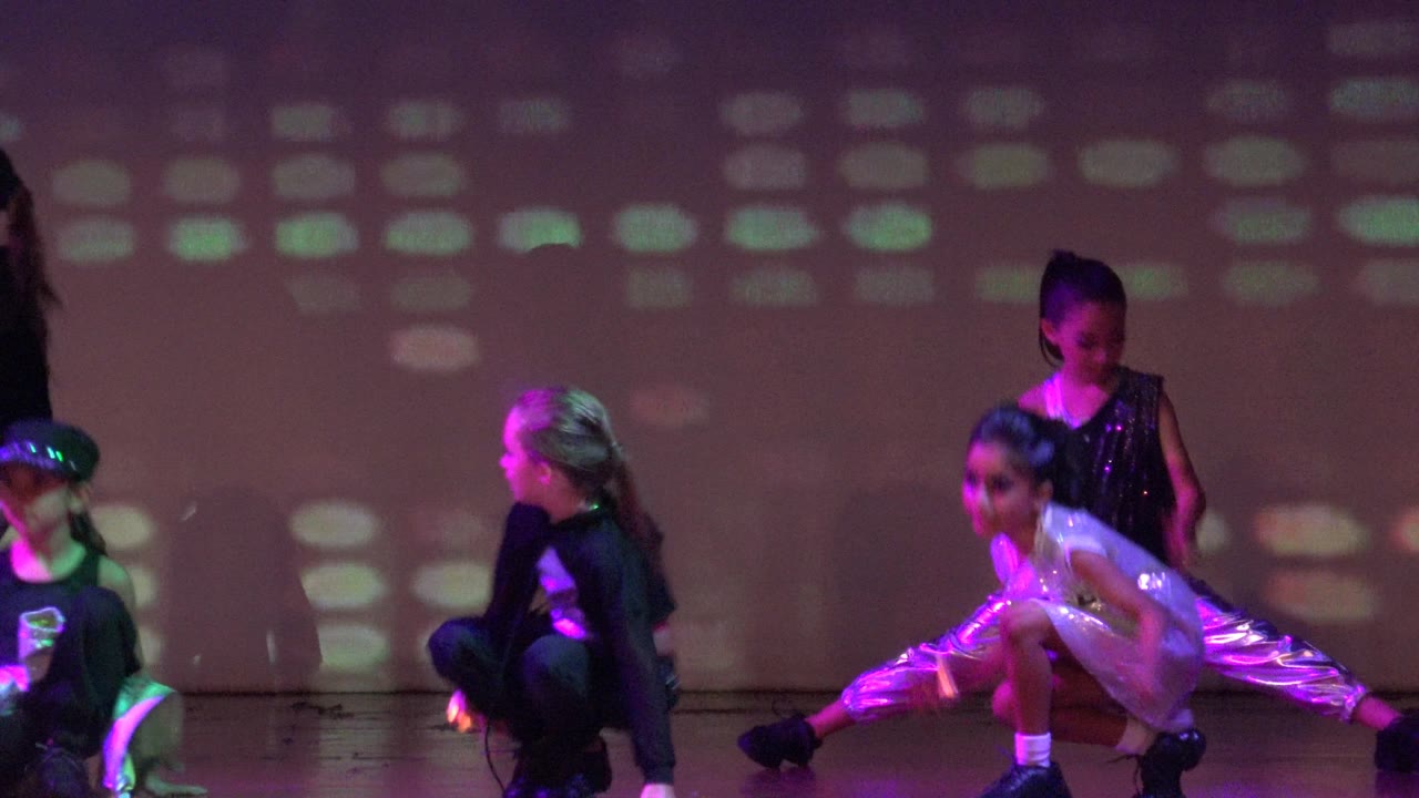 The dance group in San Jose del Cabo BCS Mexico moves into varied levels, with one child crouching and another in a split while colored light dots spread across the background.