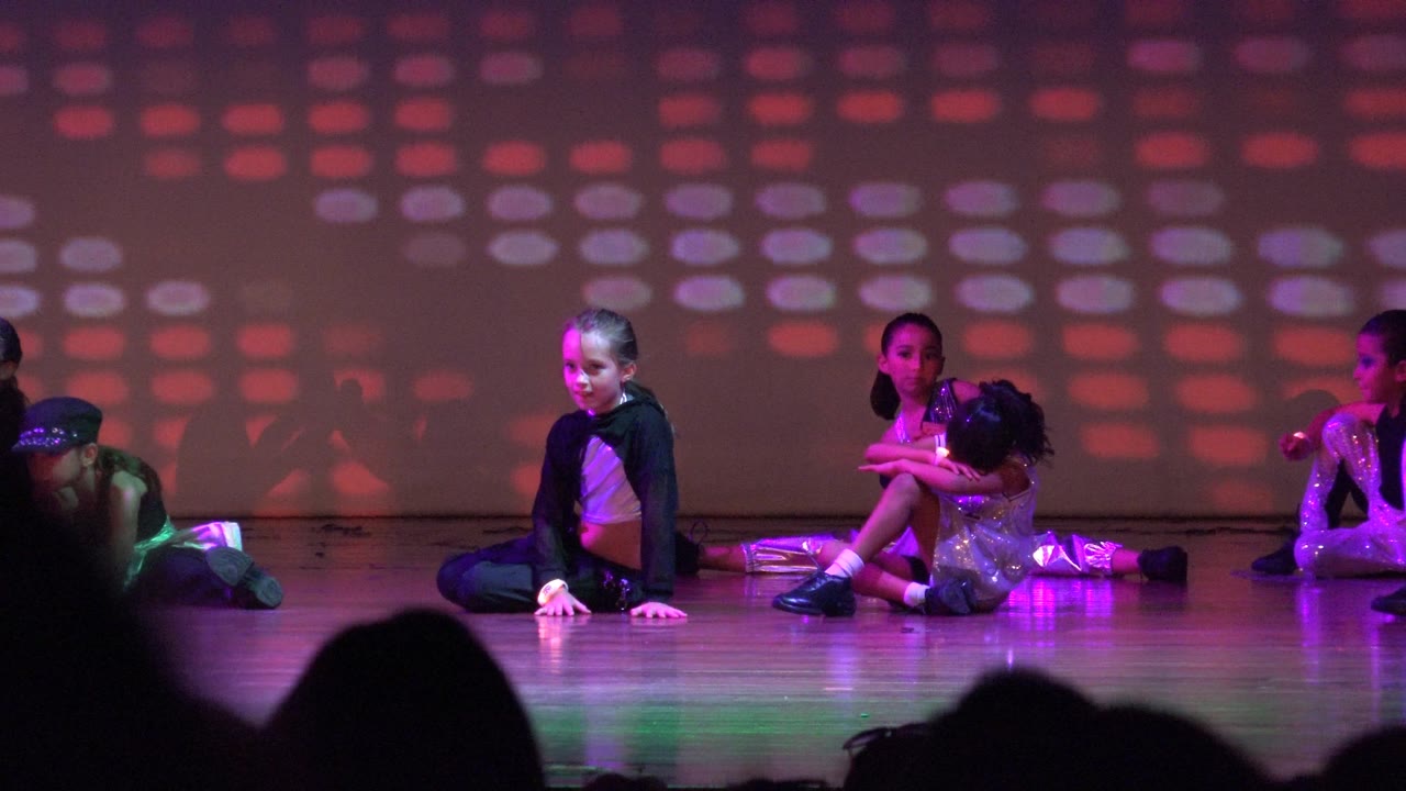 Children sit across a stage in San Jose del Cabo BCS Mexico as red and purple light patterns fill the backdrop and audience silhouettes line the bottom of the frame.
