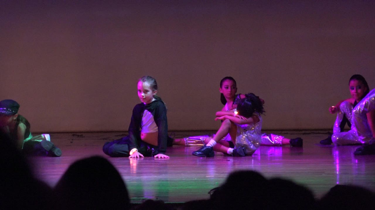 Children sit in a low stage formation in San Jose del Cabo BCS Mexico with a central performer facing forward and purple-green lighting reflected on the floor.