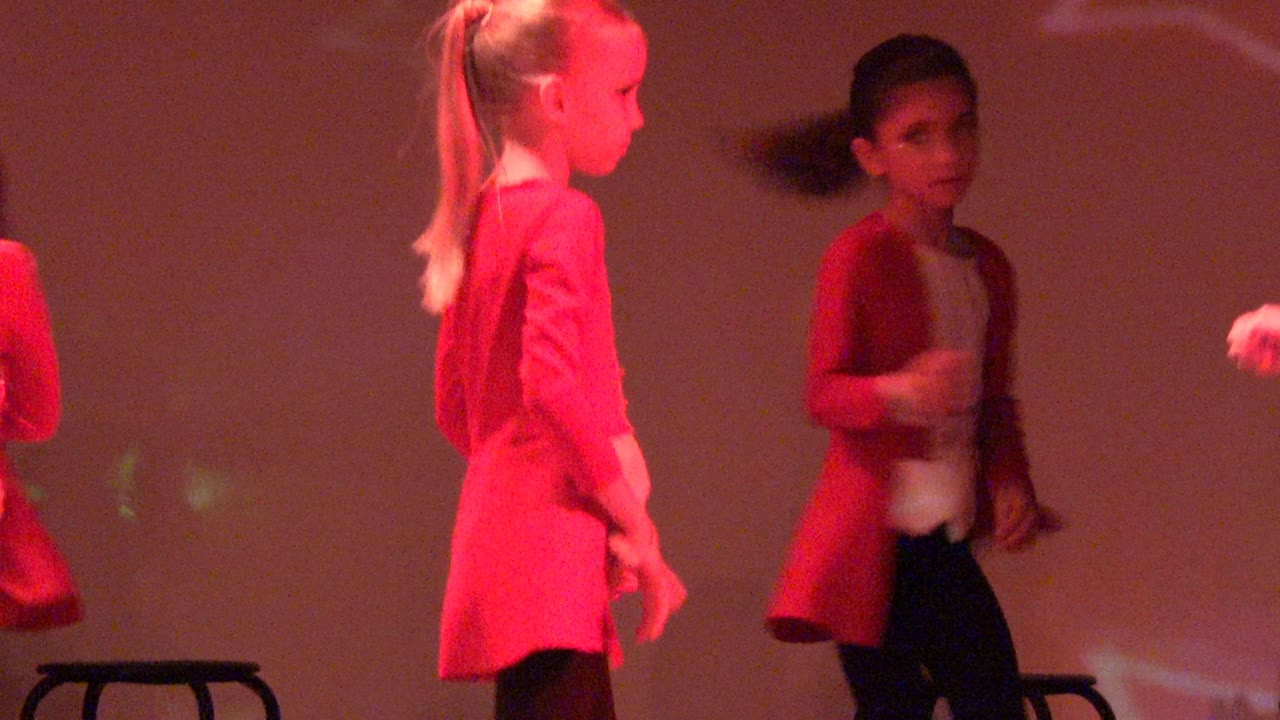 Three children in red jackets perform arm movements under red stage light.