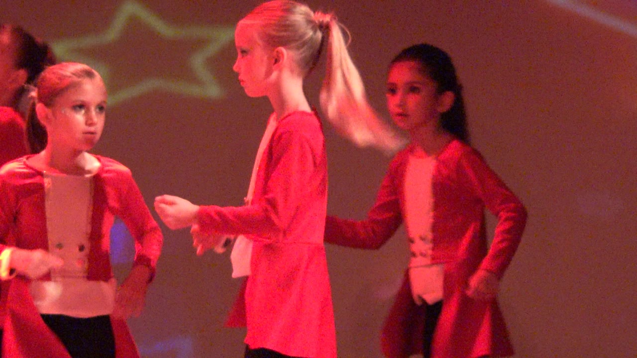 At 0:02, children in red costume jackets pass close to the camera side of the stage in San Jose Del Cabo, Mexico, with motion blur and dim star projections behind them.