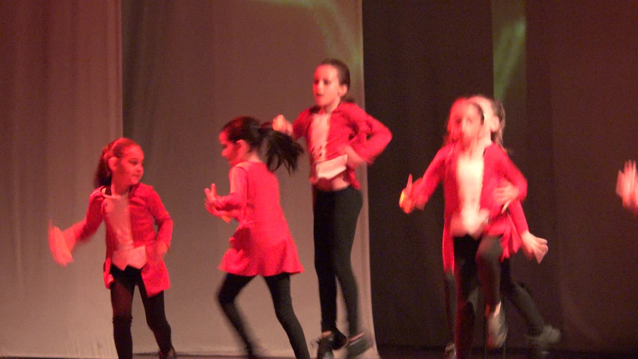 Children in red jackets and black pants perform on a stage in Mexico, with one dancer facing sideways and others partly blurred by movement.