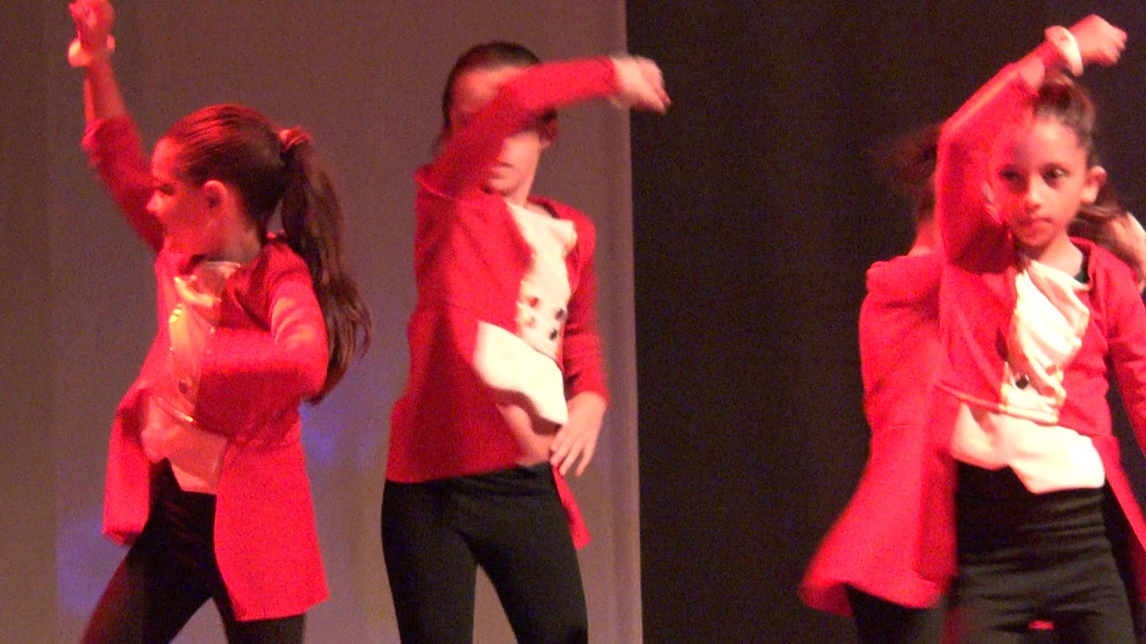 A child in a red jacket and ponytail turns in profile on the Mexico stage, with a plain backdrop and warm red wash behind her.