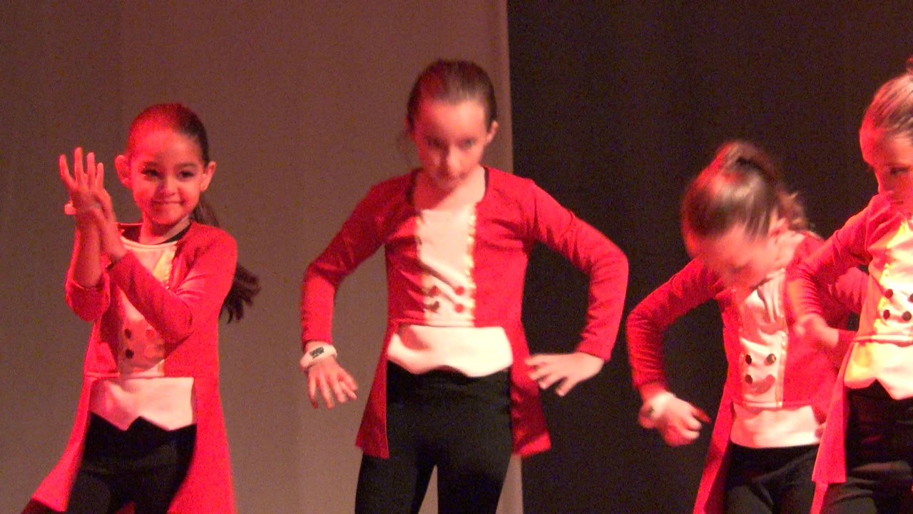 A young girl in a red and white dance costume claps near the front of an indoor stage in San Jose Del Cabo BCS Mexico, with red light on a dark backdrop.