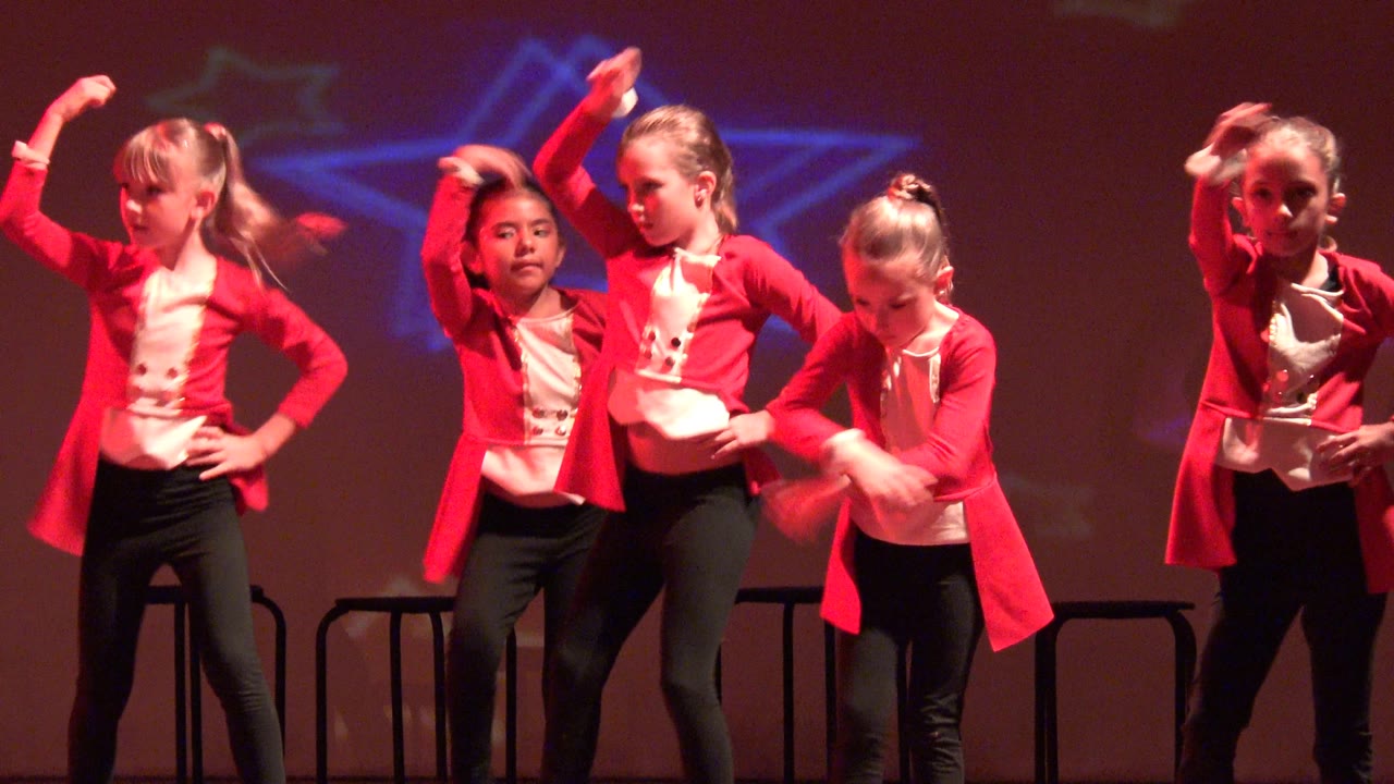 Young girls perform a stage dance routine in San Jose Del Cabo BCS Mexico, with red jackets, black pants, raised arms, and star-shaped light projections behind them.