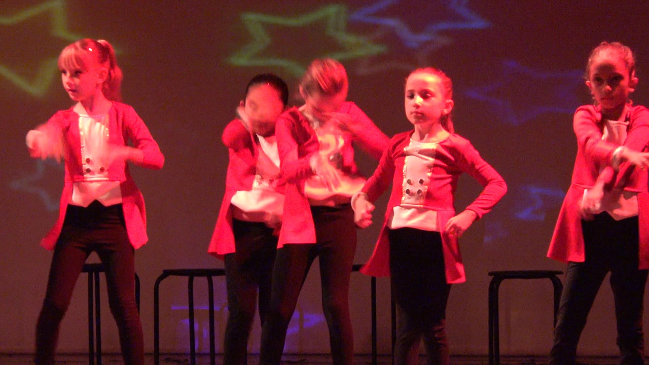 Girls in matching red jackets hold dance poses onstage.