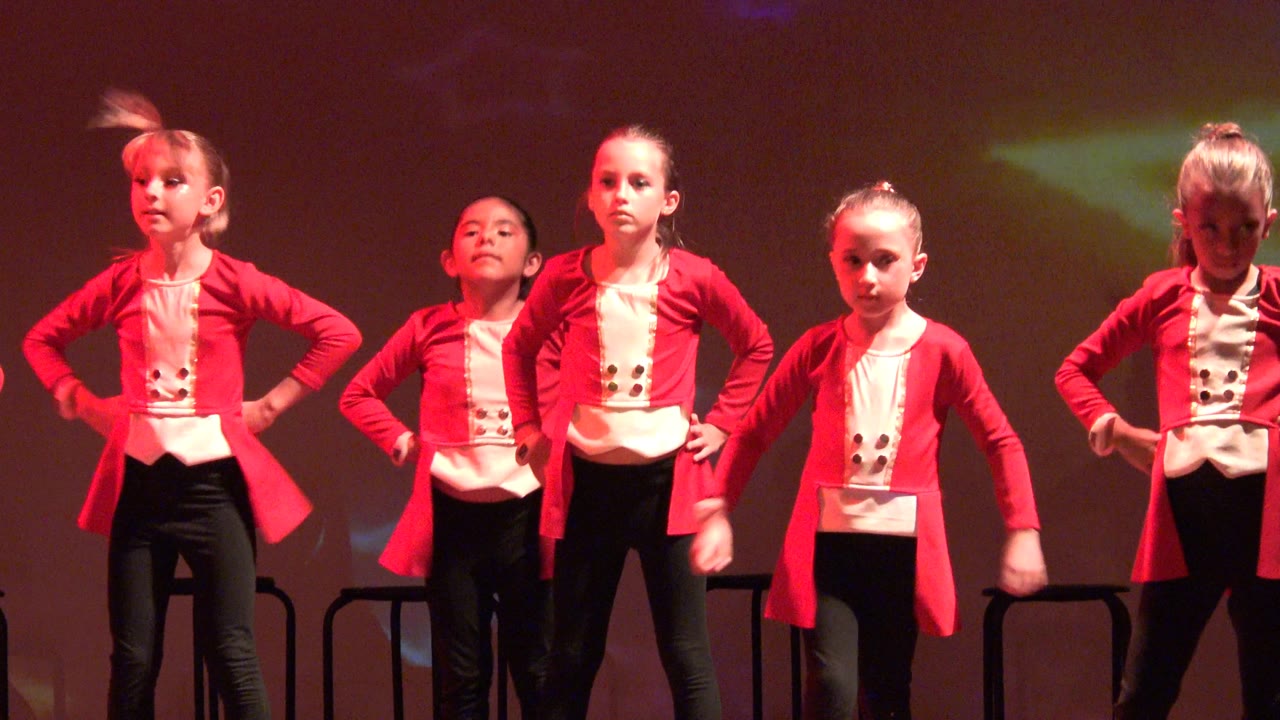 Young dancers in matching red jackets move through the routine on a stage in San Jose Del Cabo BCS Mexico, with one child blurred by arm motion and projected stars on the rear wall.