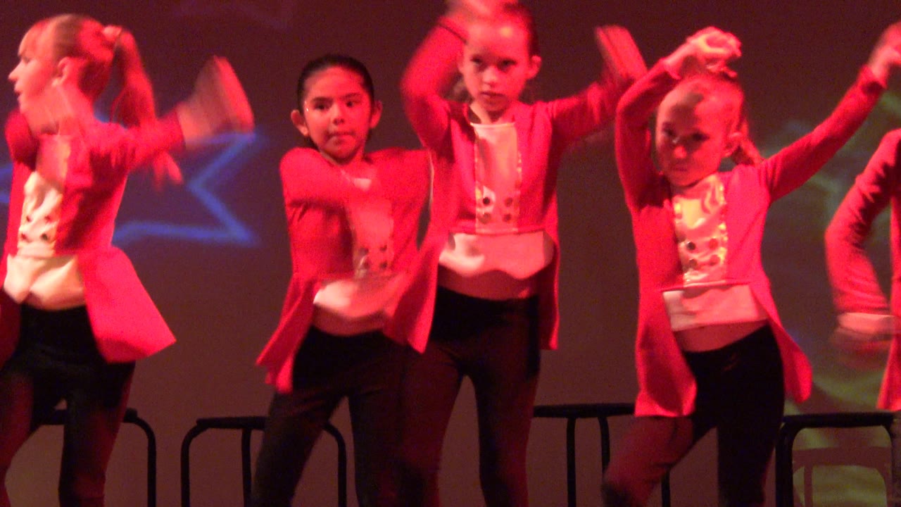 Children in red jackets continue synchronized arm movements on stage.
