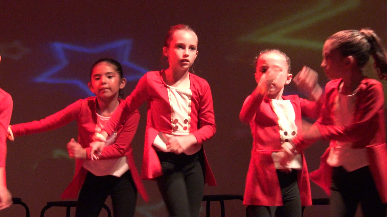 Children in red jackets and black pants perform on a stage in San Jose del Cabo BCS Mexico, with colored star projections on the backdrop and several faces visible.