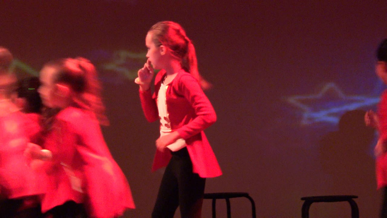 Dancers in red jackets move quickly across the stage near stools.