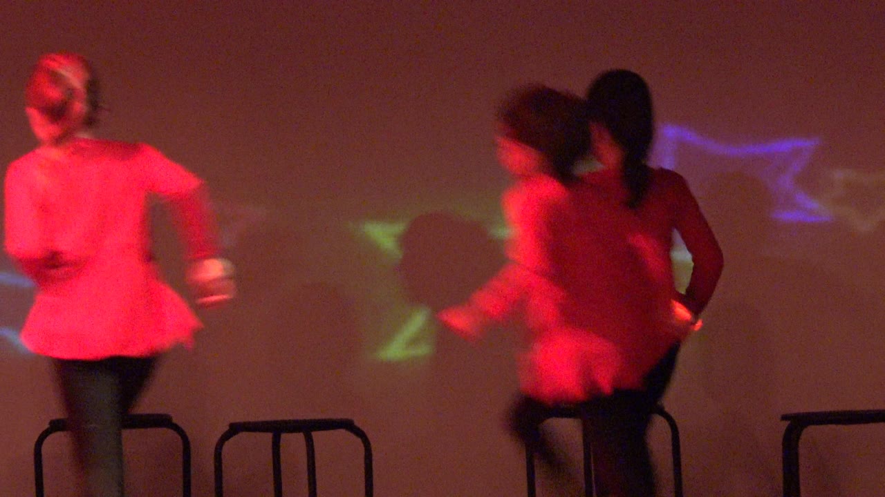 Several children in red jackets move away from the camera on a stage in San Jose del Cabo BCS Mexico, with stools lined up behind them and colored stars projected on the wall.