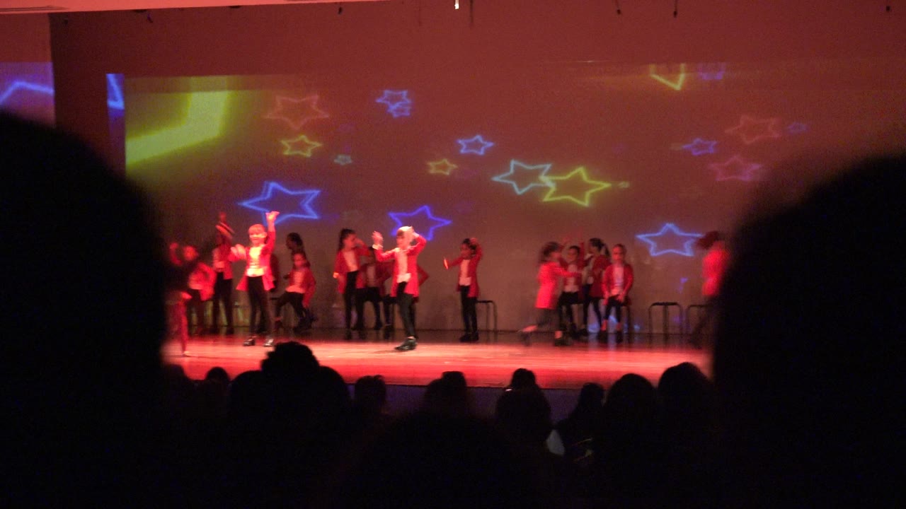 A wide San Jose del Cabo BCS Mexico stage view shows performers in red and white spread across the stage with audience heads silhouetted in the foreground and colorful star lights on the backdrop.