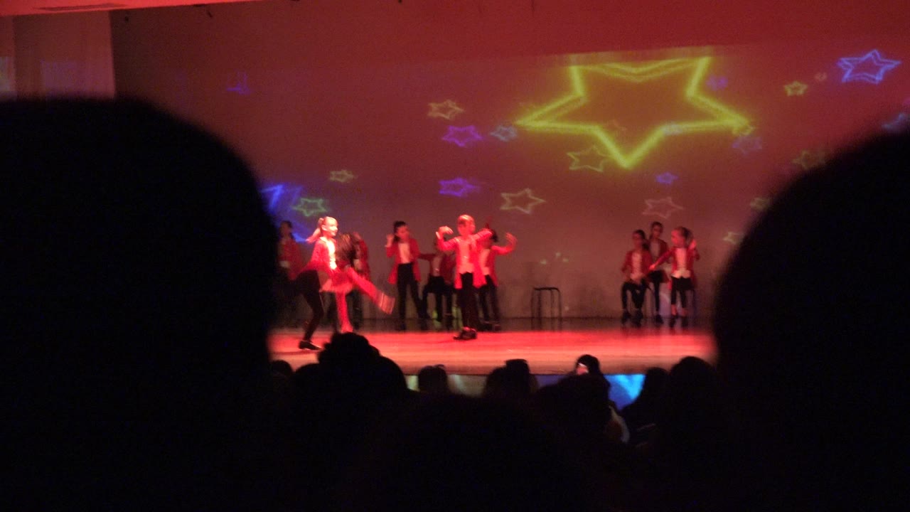Stage dancers in red and white with large audience silhouettes in foreground