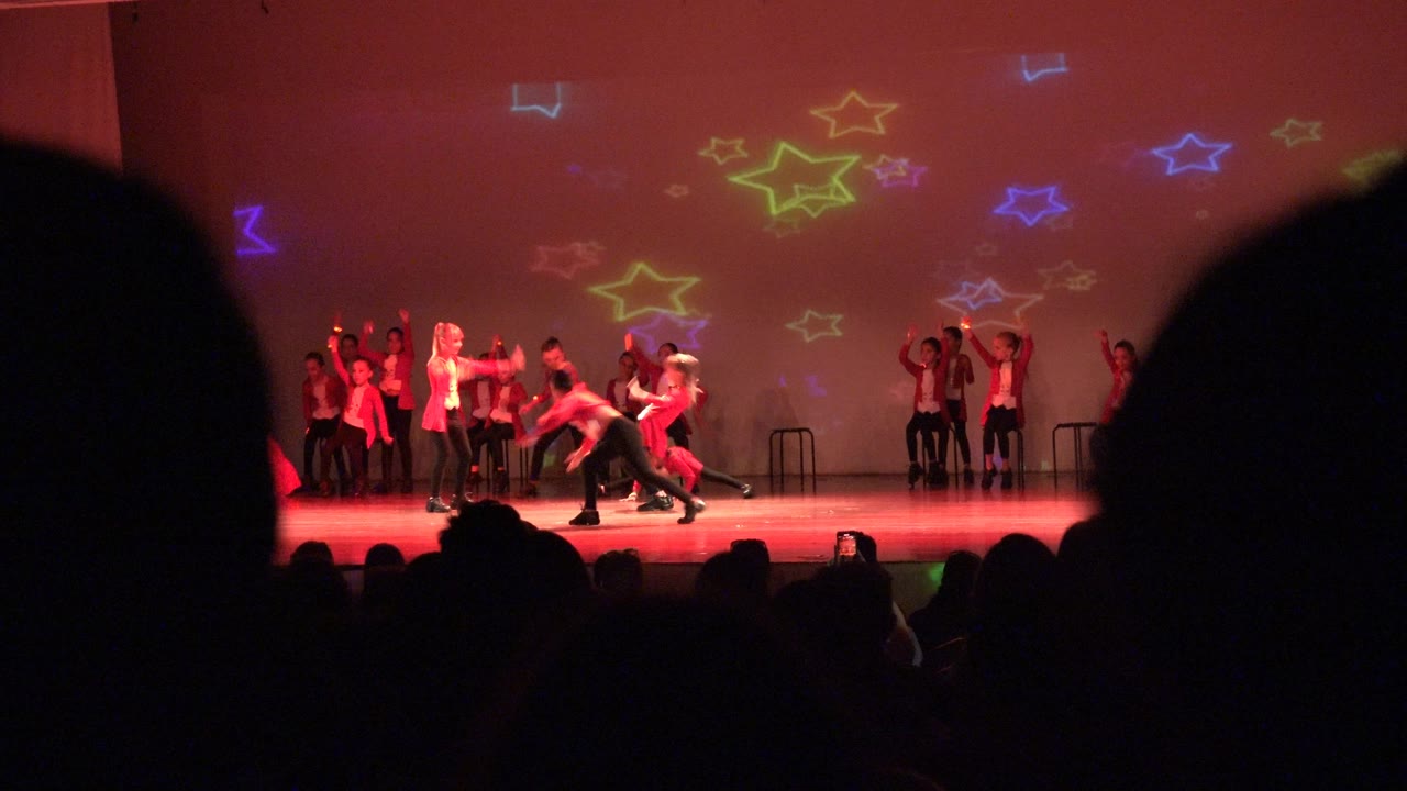 At 0:02, performers in red and white continue the San Jose del Cabo BCS Mexico routine across the stage, with colorful star projections above them and audience silhouettes along the bottom edge.