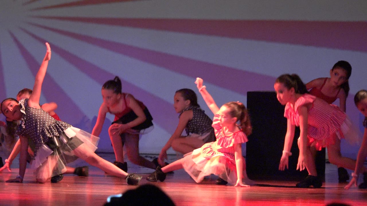 Young girls perform on a stage in San Jose del Cabo BCS Mexico, with one dancer extended sideways and others crouching or standing under red-purple light.