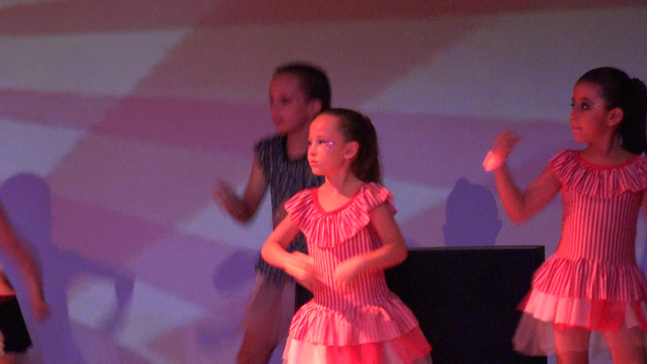 Children perform a synchronized dance routine on a stage in San Jose del Cabo BCS Mexico, with pink lighting, striped dresses, and geometric backdrop shapes.