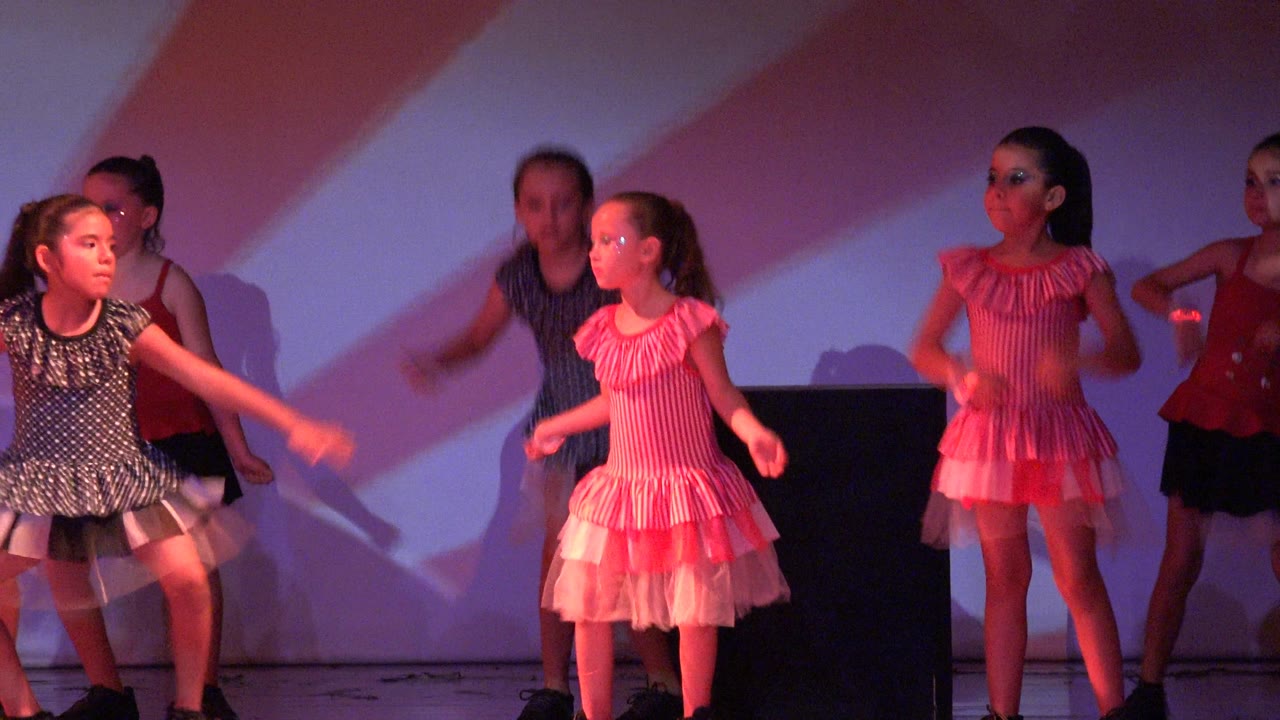 Children perform across the stage in San Jose del Cabo BCS Mexico, including a central girl in a dark patterned dress and another child in a pink striped costume.