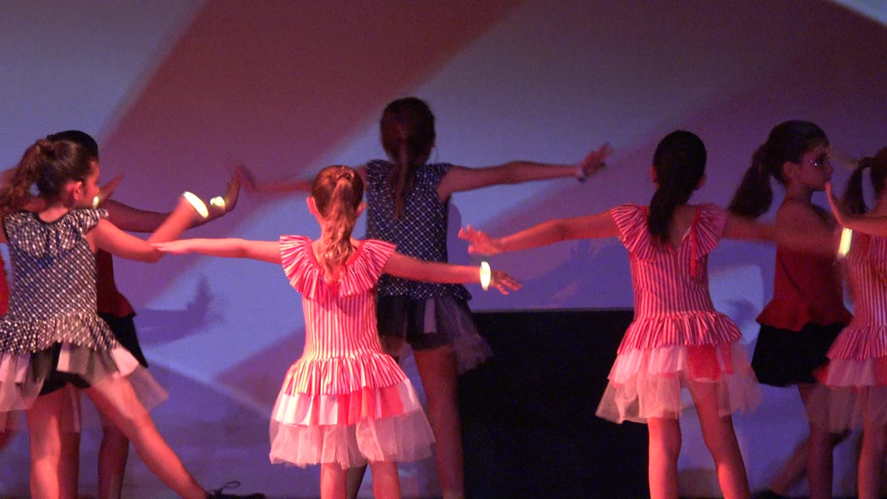 At the start of the clip in San Jose del Cabo, Mexico, young girls in colorful dresses extend their arms during a stage dance routine under red and purple lighting.