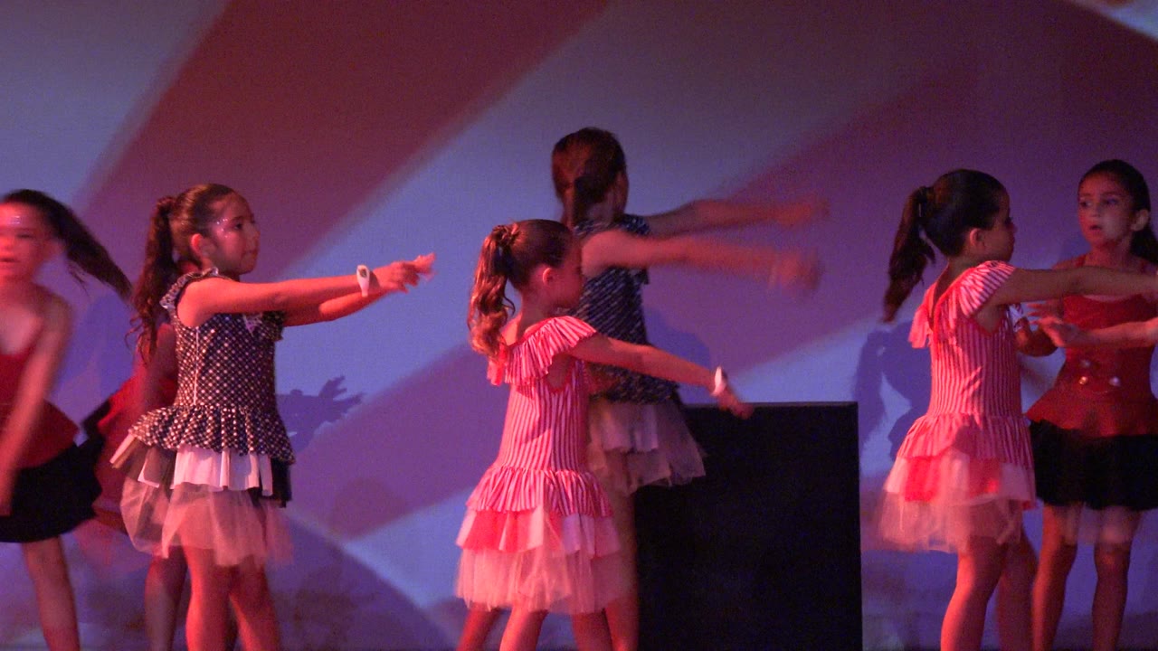 Girls continue a stage dance routine with arms extended across the frame.
